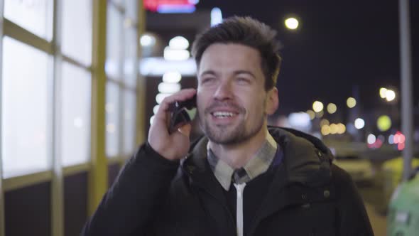 A Young Man Talks on a Smartphone with a Smile While Walking Down a Street in an Urban Area at Night