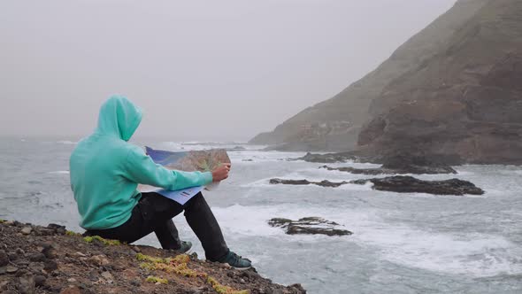 Traveler with Dog Holding Map in Front of Powerful Waves Splashing Into Rocky Coastline