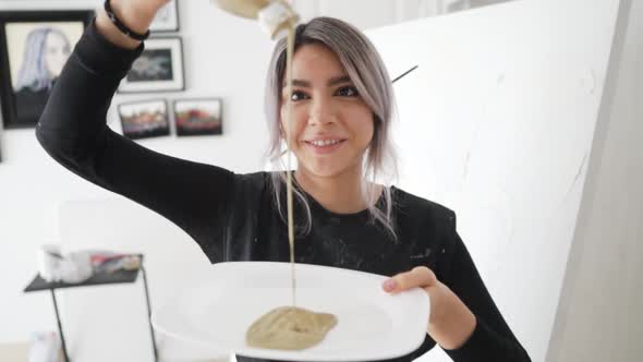 Cheerful Female Painter Pouring Paint On A White Plate Inside The Studio - medium shot, slow motion