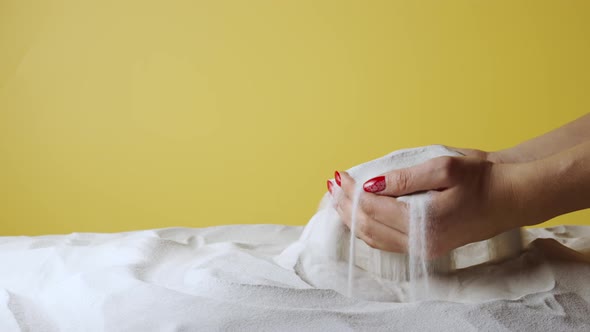 Woman Picks Up Palms Full of White Dry Sand and Pours It Out Against Yellow Background