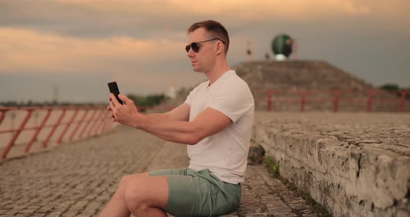 Young Man Making Selfie Wearing Sunglasses Sitting at City Embankment
