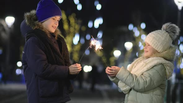 Two Cute Young Children Boy and Girl in Warm Winter Clothing Holding Burning Sparkler Fireworks on