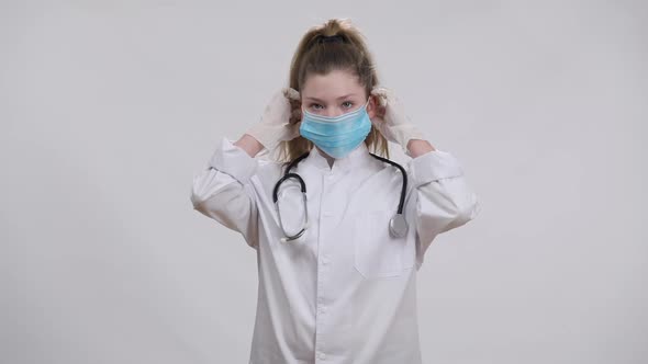 Confident Caucasian Girl in Doctor Gown Putting on Coronavirus Face Mask Crossing Hands Looking at