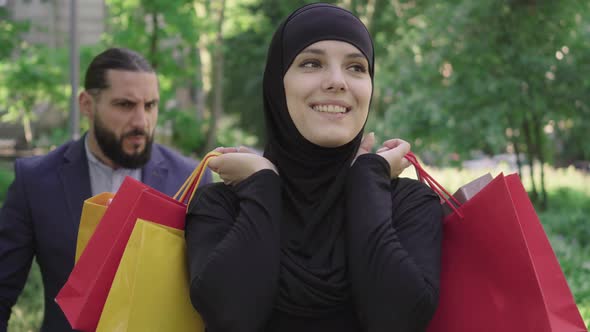 Young Satisfied Muslim Woman in Hijab Walking with Shopping Bags and Smiling As Blurred Angry Middle