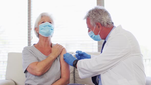 Male caucasian doctor wearing face mask giving injection to senior caucasian woman at hospital