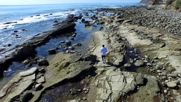 Tracking shot of a young man running on a rocky ocean beach shoreline