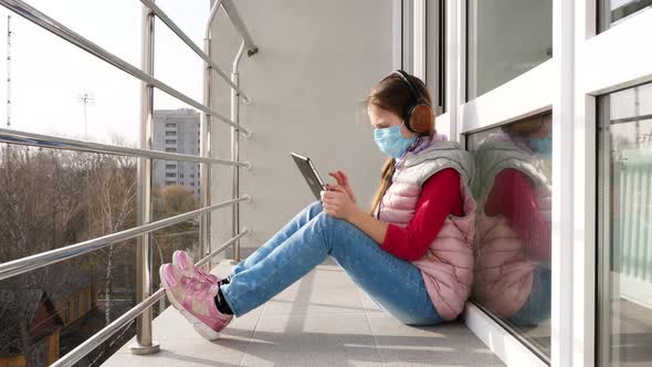 Teenager Girl in Mask and Headphones, Typing, Working on Tablet, on Open Balcony. Spring Sunny Day