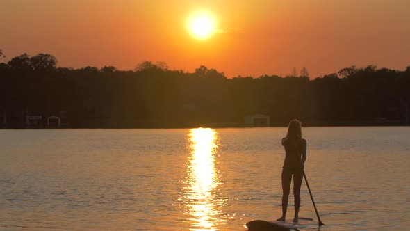 A young woman sup stand-up paddleboarding on a lake at sunset.