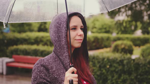 Young Happy Beautiful Woman with Transparent Umbrella in Rain Standing in City Park