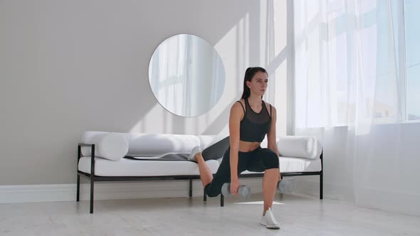 Brunette in Black Sportswear in a White Apartment Makes a Split Squat with Dumbbells in Her Hands