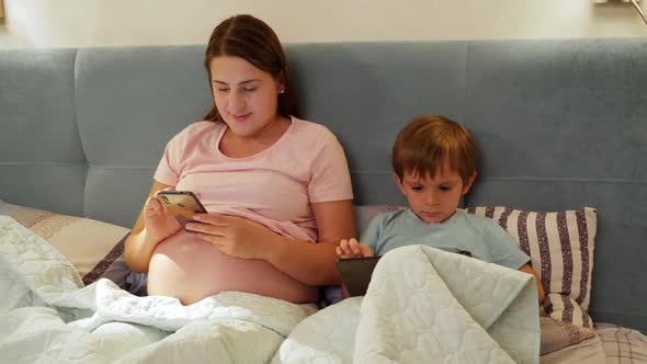 Young Pregnant Mother with Little Son Lying in Bed and Using Smartphone and Tablet Computer Before