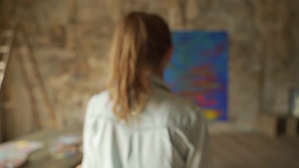 Rear View Female Artist Approaches an Easel with a Painting