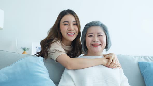 Portrait of Asian lovely family, young daughter hugging senior older mother sit on sofa in house.