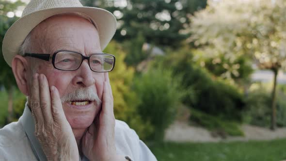 Senior Man Rubs His Head and Face From Fatigue on Bench in the Summer Park