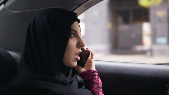 Young Attractive Muslim Woman in Hijab Sitting on the Back Seat in Taxi and Talking on the Phone