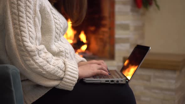 A Young Pregnant Woman is Sitting in an Armchair By the Fireplace and Typing on a Laptop