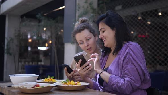 Two Cheerful Girlfriends Showing Each Other Something Funny in Their Phones During Lunch. Enjoying