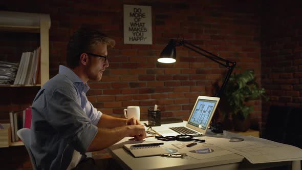 Caucasian Male with Light Beard and Blue Shirt Wearily Takes Off Glasses and Rubs Nose Near Computer