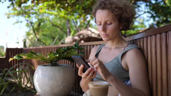 Young Busy Lady Working Remotely Through Her Smartphone From a Cafe and Drinking Plant Based Coffee