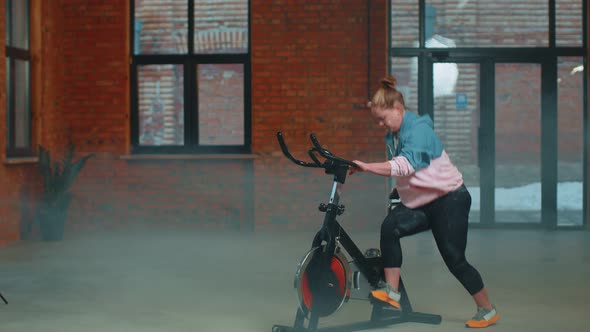 Athletic Girl Performing Aerobic Riding Training Exercises on Cycling Stationary Bike in Foggy Gym