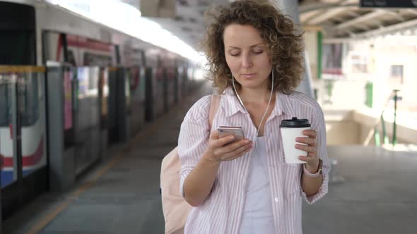 Female Passenger With Coffee Using Smart Phone On Platform Waiting For Train