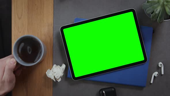 A Man Puts Cup of Tea on the Desktop Next to a Tablet with a Green Screen
