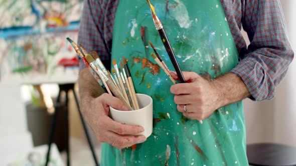 Closeup of Artist's Male Hands Holding Brushes at Level of Abdomenapron Stained with Paint