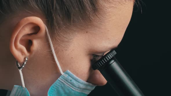 Girl Scientist Looks Into the Lens of a Microscope in a Mask Closeup