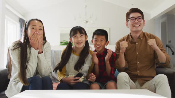 Excited asian parents watching sport on tv sitting on couch with son and daughter cheering