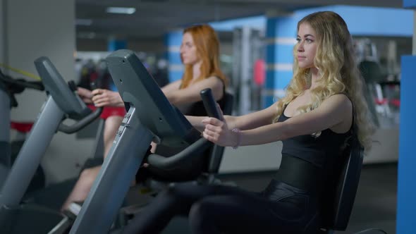Side View of Confident Fit Blond Sportswoman and Redhead Woman at Background Riding Exercise Bikes