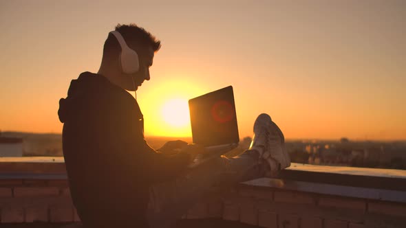 A Male Freelance Programmer Sits on a Skyscraper Roof with a Laptop and Beer Typing Code on a