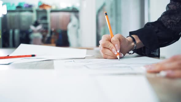 Close-up of seamstress hand with a sharp pencil draws a sketch of a stylish dress on a paper.