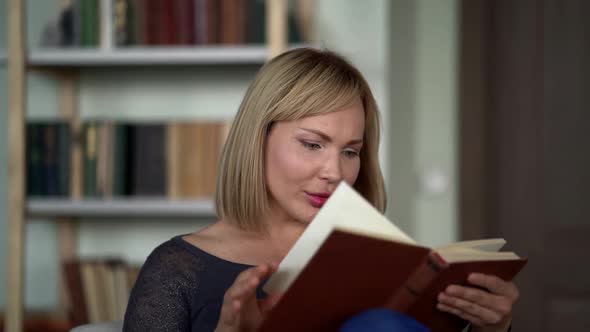 a Blonde with Bright Lipstick on Her Lips Sits Against the Background of Bookshelves and Flips