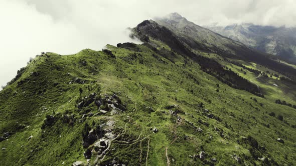 Aerial drone shot of a grass covered mountain top with a small trail leading along. More green mount