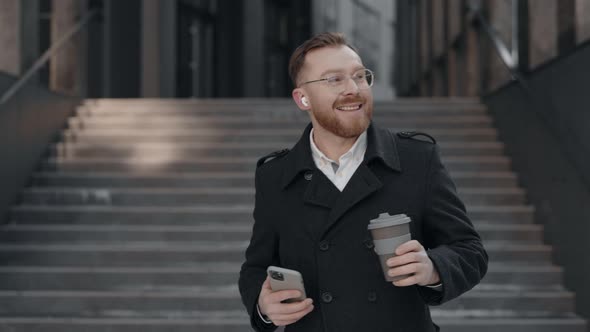 Businessman is Walking Near the Business Center with a Cup of Coffee and Smartphone in His Hand