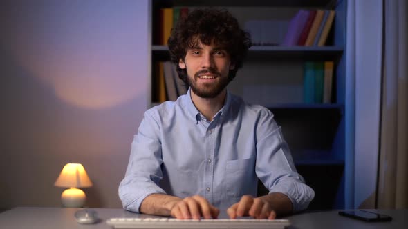 POV shot of happy curly young man looking at camera and typing online message using computer.