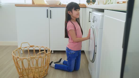 Asian young little kid doing laundry, put dirty clothes into washing machine in kitchen at house.