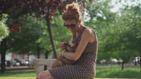 A Mature Woman Sits on a Bench in a City Park with a Dachshund Dog Holding It in Her Arms and