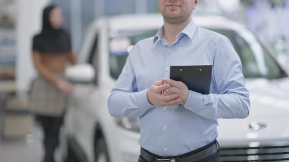Unrecognizable Car Dealer Smiling Standing in Showroom with Blurred Woman in Hijab at Background