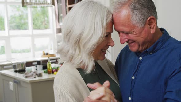 Romantic senior caucasian couple at home dancing in the kitchen and smiling