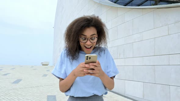 Excited Joyful African American Young Curly Woman with Glasses Walking Outdoors Holding and Using