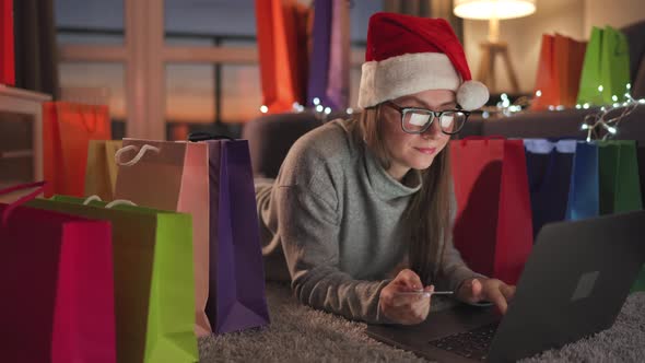 Woman with Glasses Wearing a Santa Claus Hat is Lying on the Carpet and Makes an Online Purchase