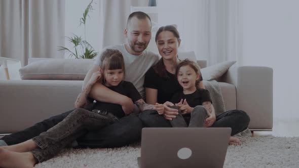 Young Family of Four with a Laptop Near Couch. Dad, Mom and Two Sister Daughters in Black T-shirts