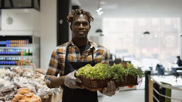 Male Smiling Worker at the Store Walks with Basket Full of Greens