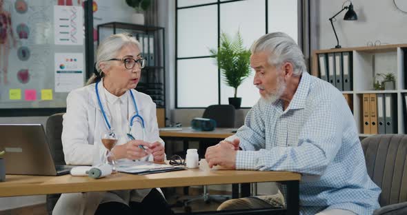 Doctor Sitting at Workplace Holding Clipboard while Advice with Senior Patient During Medical Visit