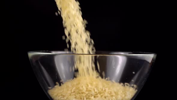 Rice is poured into a glass bowl on a black background. Slow motion.