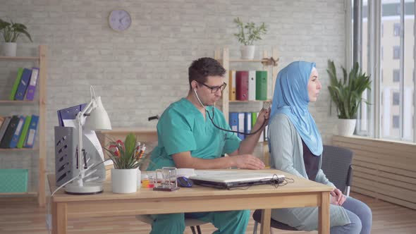 Young Male Doctor Using a Stethoscope Listens To a Young Muslim Woman in a Hijab