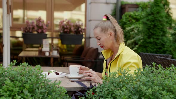 a Blonde in Red and Yellow Clothes Sits with a Phone and a Cup of Coffee at a Cafe Table Against a