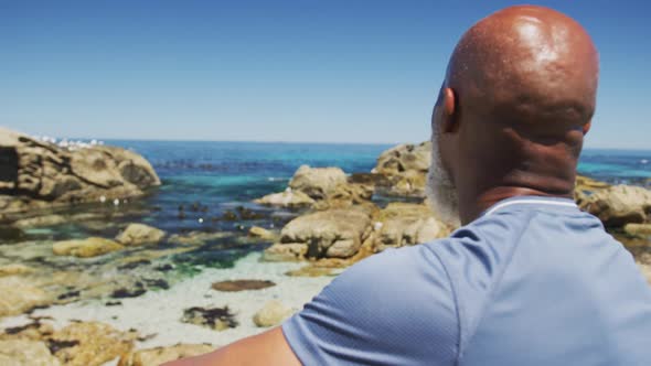 Portrait of smiling senior african american man exercising sitting on rocks by the sea