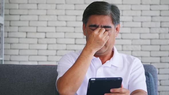 Senior man is suffering from eye strain due to his long-time looking at the digital tablet.